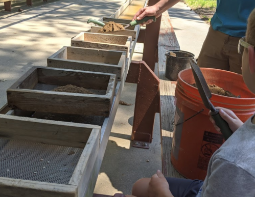 A person and a child use wooden boxes to sift sand, with tools and a bucket nearby on a sunny day.