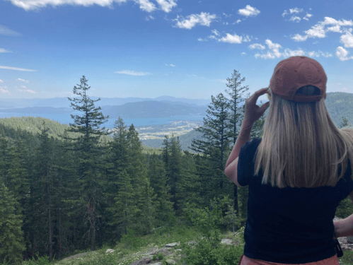 A person with long hair and a cap stands among trees, gazing at a scenic view of mountains and a lake.