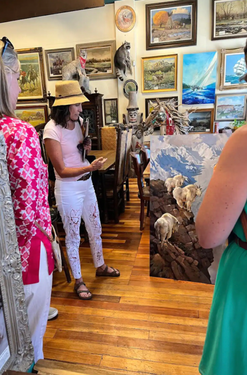 A woman in a hat discusses a painting of mountain goats in an art-filled room with two other people.