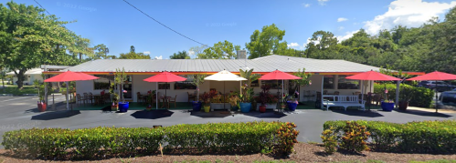 A colorful outdoor café with red umbrellas, surrounded by greenery and a clear blue sky.