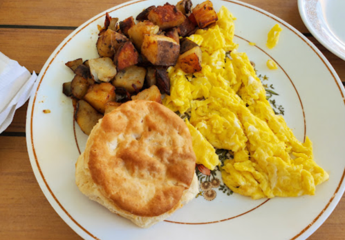 A plate with scrambled eggs, a biscuit, and seasoned roasted potatoes on a wooden table.