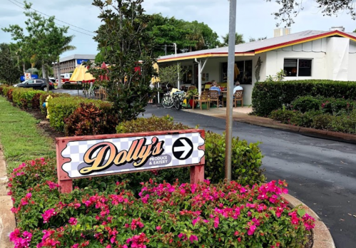 Sign for Dolly's Produce & Eatery with colorful flowers in the foreground and outdoor seating visible in the background.
