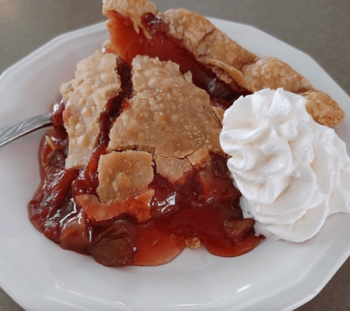 A slice of cherry pie with a flaky crust, served with a dollop of whipped cream on a white plate.