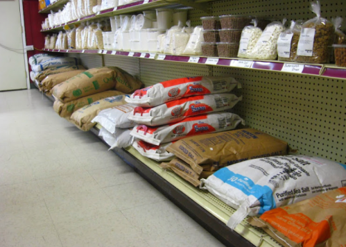 A grocery store aisle displaying various bags of grains and salt on shelves, with containers of bulk items above.