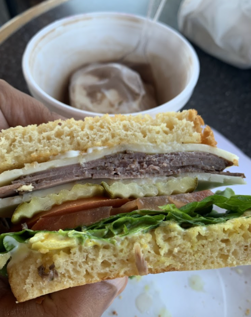 A close-up of a hand holding a sandwich with layers of meat, pickles, tomato, lettuce, and mayonnaise, with a dessert in the background.