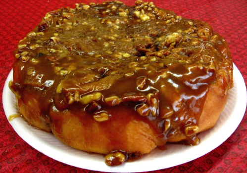 A large, sticky cinnamon roll topped with a thick layer of caramel and chopped nuts on a white paper plate.