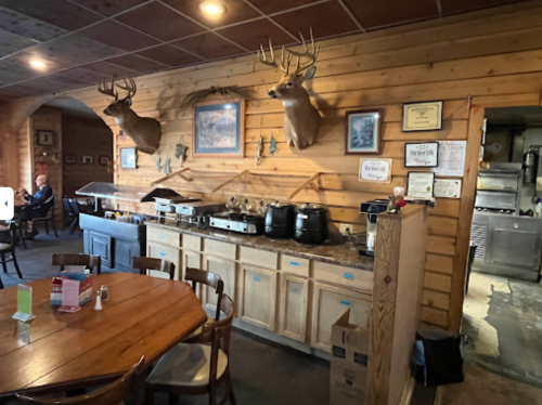 A rustic restaurant interior with wooden walls, a round table, and deer heads mounted on the wall.