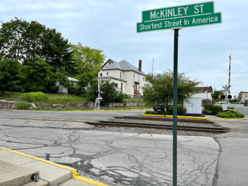 Sign for McKinley St, labeled as the shortest street in America, with a house and railroad tracks in the background.