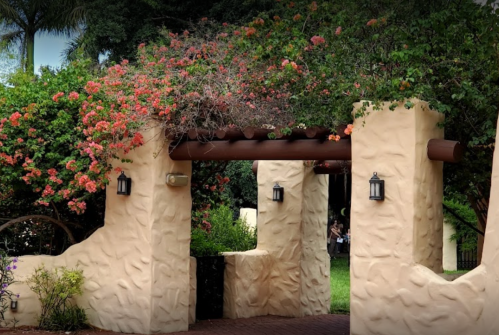 A textured stucco archway adorned with vibrant flowers and greenery, leading into a lush garden area.
