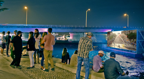 People gather by a river at night, enjoying a vibrant scene with a bridge and a large screen displaying a video.