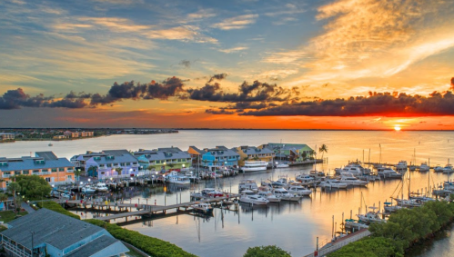 Colorful waterfront buildings reflect in calm waters at sunset, with boats docked in the marina under a vibrant sky.