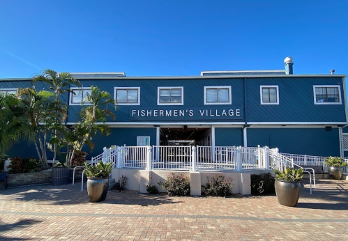 A blue building with the sign "Fishermen's Village," featuring a patio and potted plants under a clear blue sky.