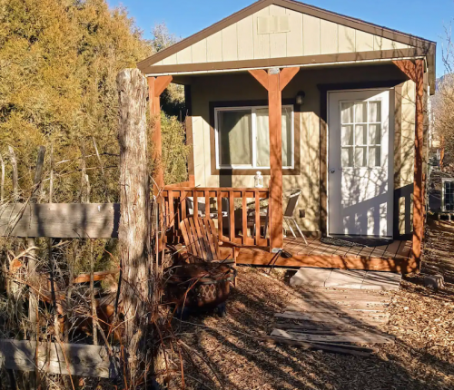 A small cabin with a porch, surrounded by trees and wooden fencing, on a sunny day.