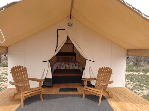 A cozy glamping tent with a bed inside and two wooden chairs on a wooden deck.