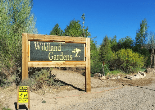 Wooden sign for Wildland Gardens surrounded by greenery, with a caution sign to slow down for kids at play.