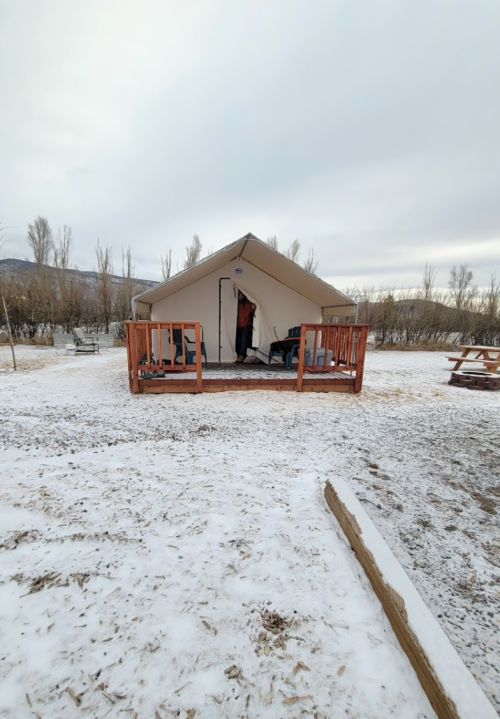 A large tent on a snowy ground, with a wooden deck and picnic tables nearby, surrounded by bare trees.