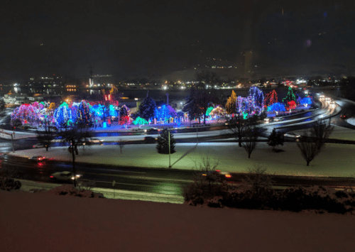 Colorful holiday lights illuminate a snowy landscape, with trees and decorations visible along a circular road at night.