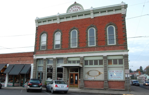 Historic brick building with large windows, "C. Young 1892" sign, and a "For Sale" sign in front. Cars parked nearby.