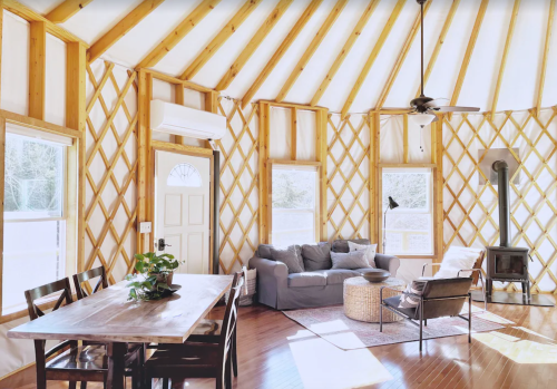 Cozy interior of a yurt with wooden beams, a dining table, sofa, and a small wood stove. Bright natural light.