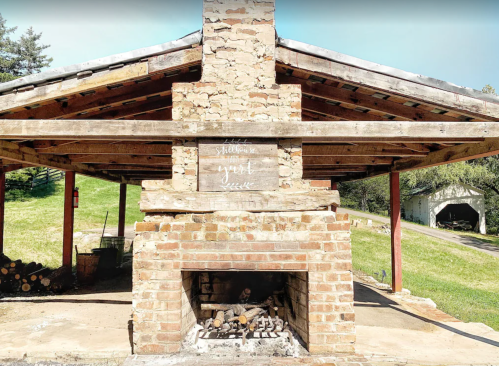 A rustic outdoor fireplace with a stone chimney, surrounded by wooden beams and a grassy landscape.