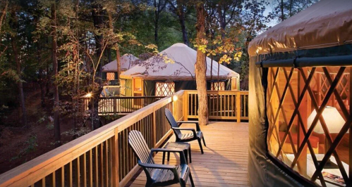 A wooden deck with two chairs overlooks cozy yurts nestled among trees at dusk.