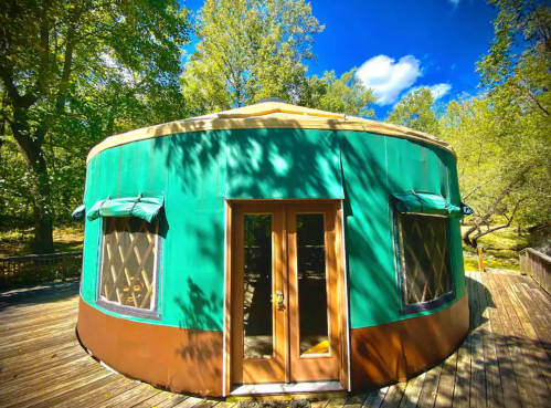 A round, green yurt with wooden doors and windows, surrounded by trees and a blue sky.