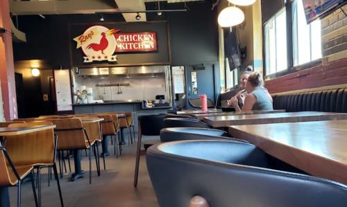 A nearly empty restaurant with wooden tables and chairs, featuring a sign for "Ray's Chicken Kitchen" in the background.