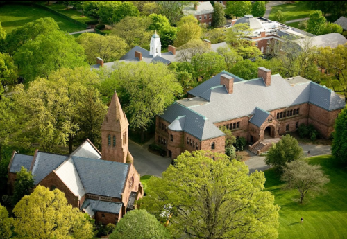 Aerial view of a campus with brick buildings surrounded by lush green trees and a clear blue sky.