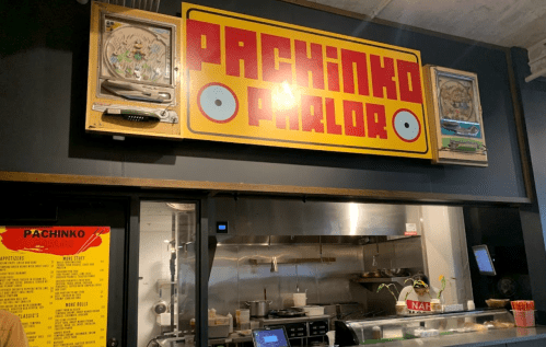 Sign for "Pachinko Parlor" above a kitchen area, featuring vintage pachinko machines and a menu on the wall.