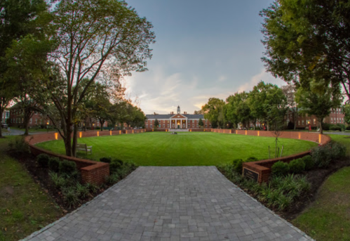 A wide view of a grassy courtyard surrounded by trees and brick pathways, with a building in the background at dusk.