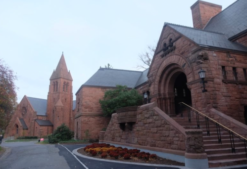 Historic stone building with a tall tower, steps leading to an arched entrance, and landscaped gardens.