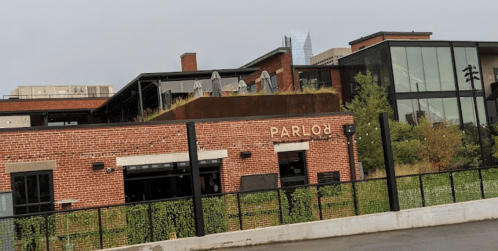 A brick building with a sign reading "PARLO" and a rooftop patio, surrounded by greenery and modern architecture.