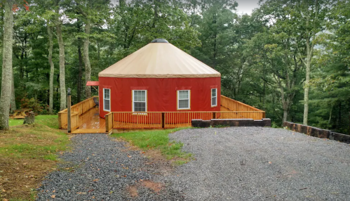 A red yurt with a tan roof, surrounded by trees, featuring a wooden deck and gravel pathway.