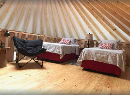 Cozy interior of a yurt with two beds, a chair, and wooden walls and floor. Natural light fills the space.