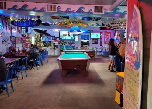 A lively bar interior with a pool table, colorful decor, and patrons enjoying drinks and conversation.