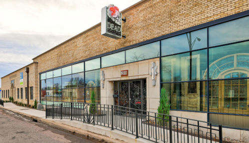 Exterior view of a brick building with large windows and a sign, featuring a modern entrance and landscaped surroundings.