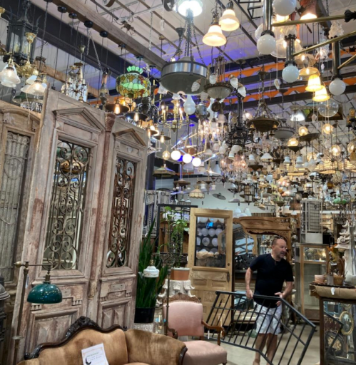 A man carries a metal frame in a store filled with various hanging lights and vintage furniture.
