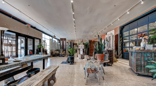 A spacious interior filled with rustic furniture, plants, and decorative items, featuring large windows and warm lighting.