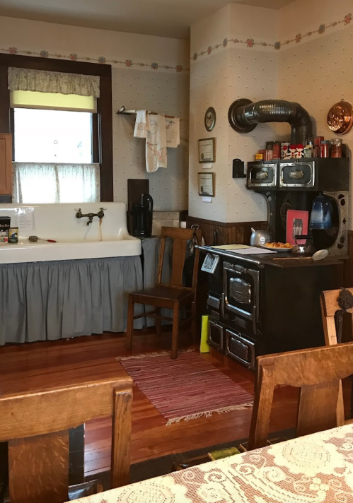 A vintage kitchen featuring a black stove, wooden chairs, a sink, and rustic decor with floral wallpaper.