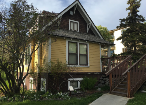 A charming yellow house with a sloped roof, surrounded by greenery and a wooden staircase leading to the entrance.
