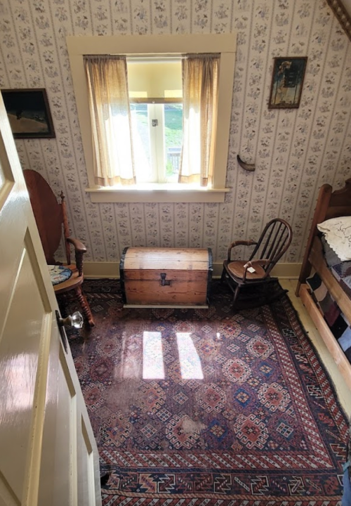 Cozy room with a wooden chest, rocking chair, and patterned rug, illuminated by sunlight through a window.