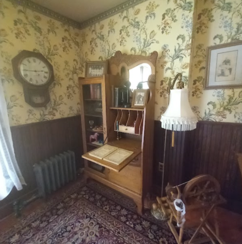 A vintage room with floral wallpaper, a clock, a wooden desk, a lamp, and a spinning wheel, creating a cozy atmosphere.