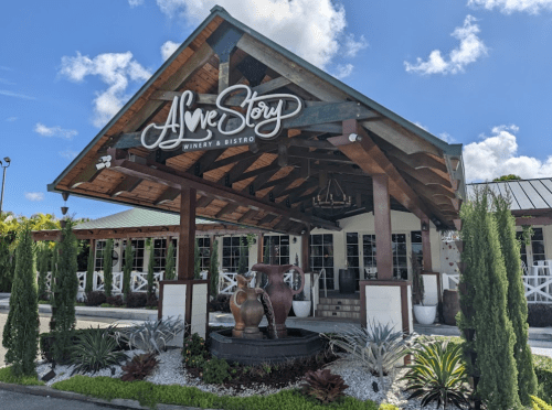 Exterior of a winery and bistro with a wooden roof, decorative pots, and lush landscaping under a blue sky.