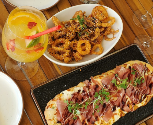 A colorful cocktail, crispy fried snacks, and a flatbread topped with prosciutto and microgreens on a wooden table.