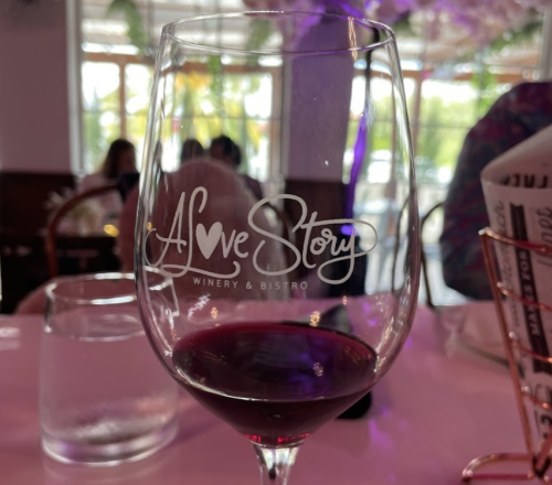 A wine glass with "Love Story Winery & Bistro" etched on it, filled with red wine, on a table setting.