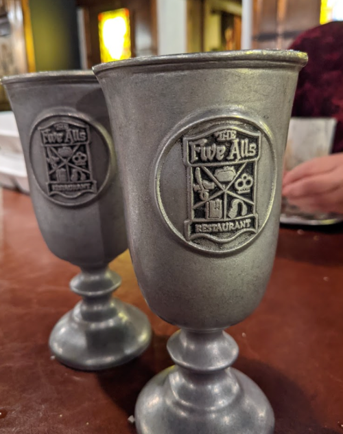 Two metal goblets with the logo of "The Five Alls Restaurant" on the front, set on a wooden table.