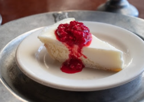 A slice of creamy cheesecake topped with raspberry sauce on a small white plate.