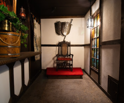 A medieval-style room featuring a throne on a red carpet, decorative shields, and rustic wooden accents.