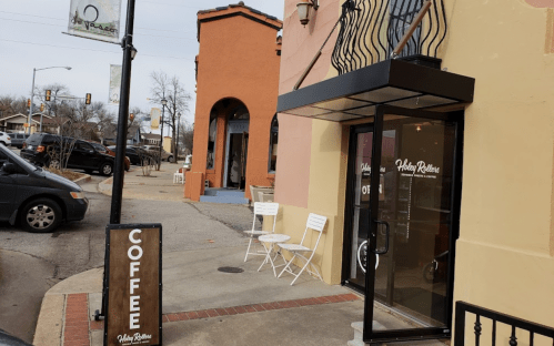 A coffee shop entrance with a sign, outdoor seating, and a car parked nearby on a cloudy day.