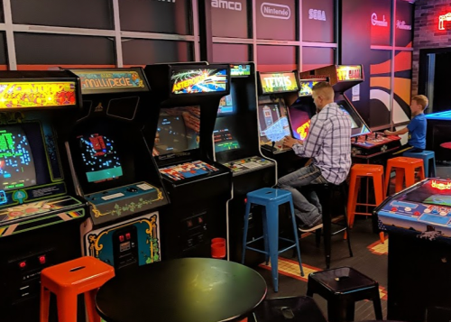 Two people play retro arcade games in a colorful gaming space filled with classic machines and bright screens.